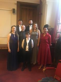Musical Director Antony Brannick flanked by soloists Rebecca Moon (Royalty, soprano) and Elspeth Marrow (Tyranny, mezzo-soprano), next to Narrator Claire Benedict (Time); behind, Jean-Pascal Heynemand (Hermes, tenor) and James Fisher (Zeus, bass); in front, Zachary Smith (Herakles, treble). 